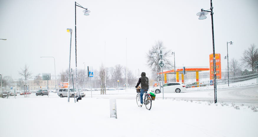 En person cyklar i snöoväder bort från kameran. I bakgrunden syns en rondell och en bensinmack.