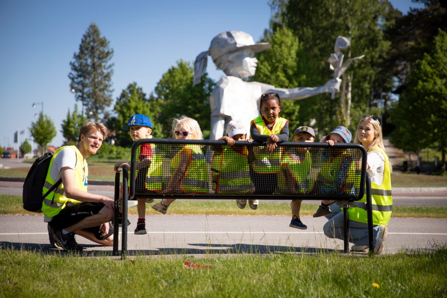 Några barn i gula västar sitter på en bänk i stadsmiljö. Intill sitter två unga vuxna på huk. De har också gula reflexvästar. 