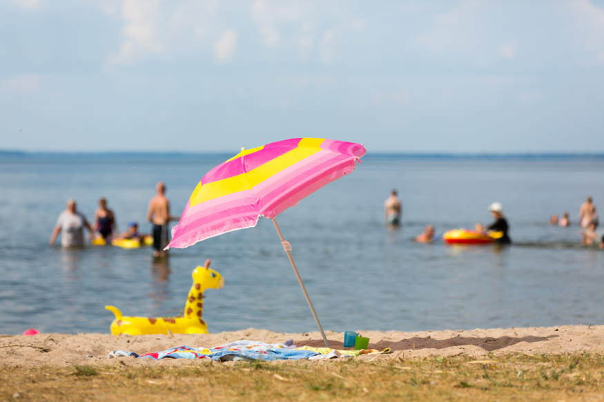 En strand där man ser människor bada. På stranden finns ett rosa och gult parasoll samt flera leksaker i sanden.