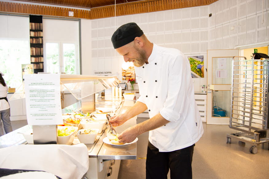 En kock/kökcchef serverar mat i en skolmatsal.