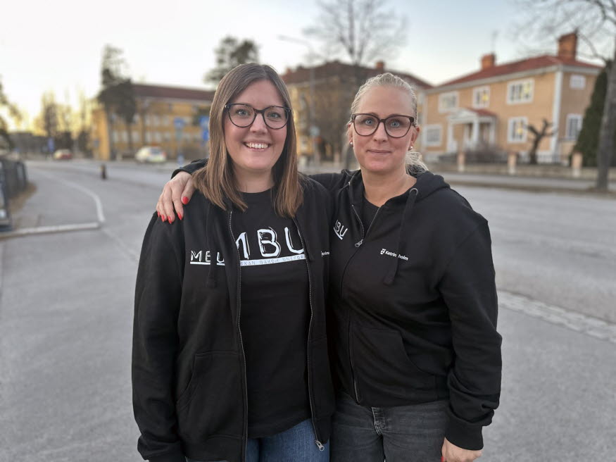 Två kvinnor står på en trottoar och tittar mot kameran. Bakom kvinnorna syns gula och orangea hus
