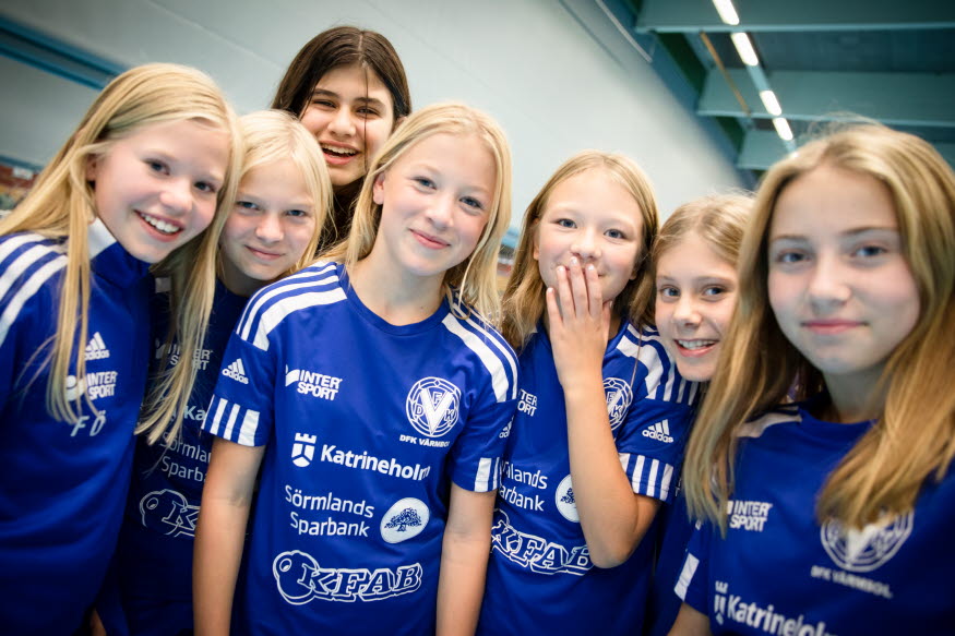 Sju unga tjejer i blå fotbollströjor, i en sporthall. 