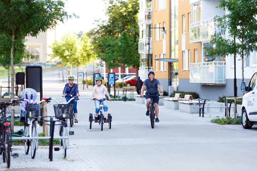 Tre personer cyklar i centrala Katrineholm. Alla tre bär hjälm. 