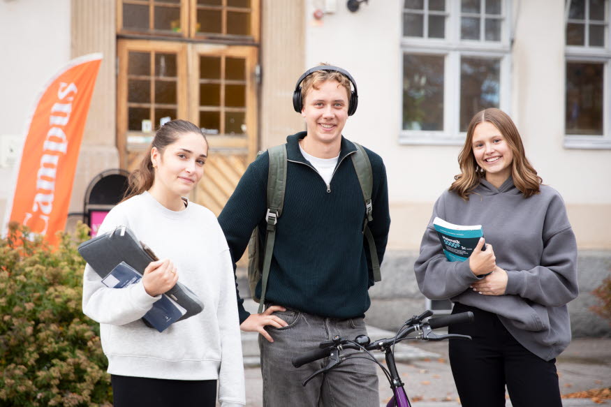 Tre unga studenter framför en vit byggnad. 