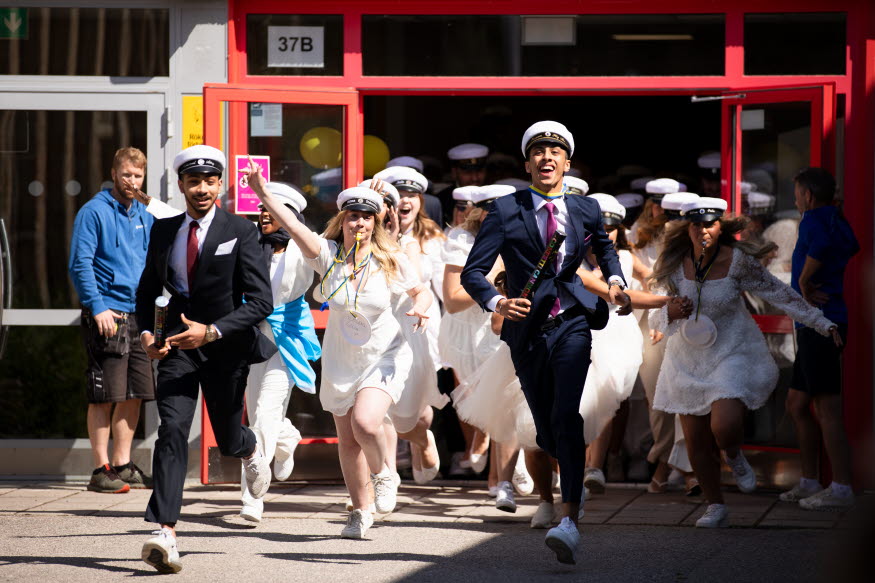 Studenter i vita klänningar och mörka kostymer springer ut genom en dörr.