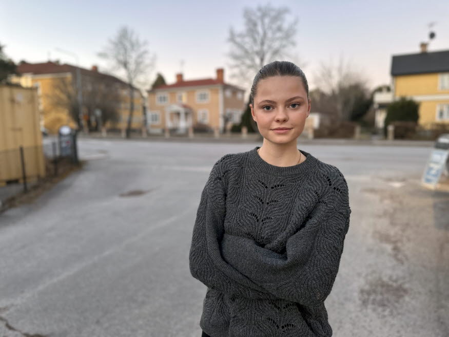 En tjej står på en väg med armarna i kors och tittar mot kameran. Bakom tjejen syns gula hus och byggnader.