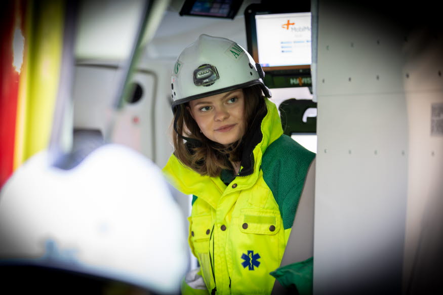 Evelina sitter i en ambulans med gul ambulansjacka och vit hjälm.