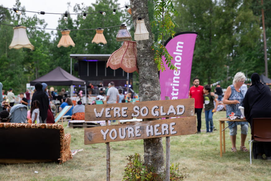 Gatufest i Nävertorp. På gräsplanen befinner sig människor i olika åldrar som tittar på scenen, äter mat och deltar i aktiviteter. Området är pyntat med lampor, vimplar och skyltar i olika färger.