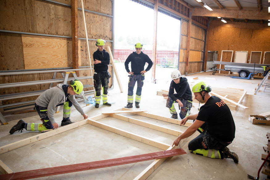 Unga personer i snickarkläder i en bygghall. 