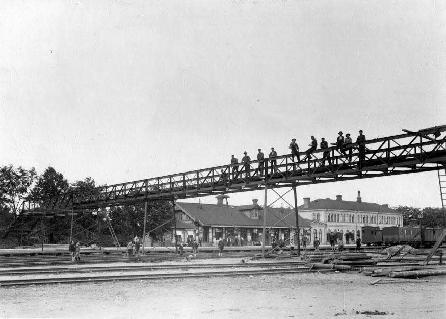 Svartvit bild som visar Järnvägsstationen och gångbron "Himlastegen". Gångbron går över järnvägsräls och på bron står människor och tittar ut.