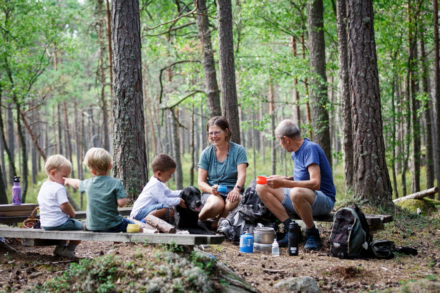 Flera personer, gamla som vuxna, och en hund sitter i en skog och fikar.
