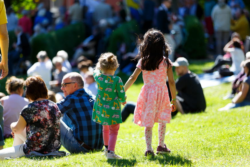 Två barn går på en gräsmatta och håller handen. I bakgrunden syns människor som sitter på gräsmattan. 