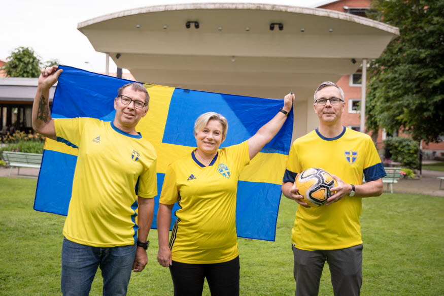 Tre personer i blågula tröjor håller i en Sverigeflagga och en boll. De står framför en ljus stor scen i Stadsparken.