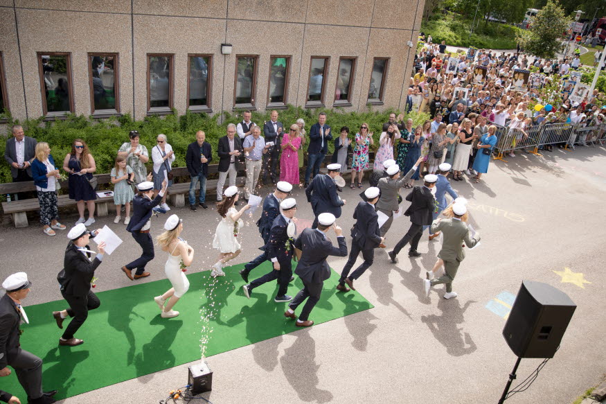 Festklädda studenter i vita mössor sprunger ut genom en byggnad. I bakgrunden mycket folk. 
