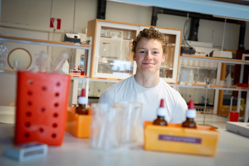 En glad person i ett klassrum med glasrör och andra experimentprylar.